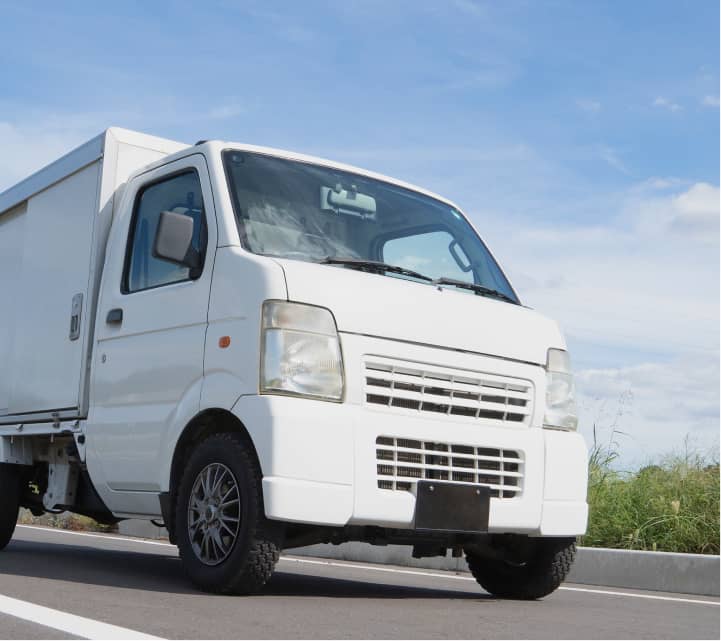 白い軽トラックが青空の下、道路を走行している配送車両